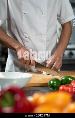 Hands of young male chef of restaurant chopping fresh vegetables on wooden board while cooking vegetarian lunch by table in the kitchen Stock Photo