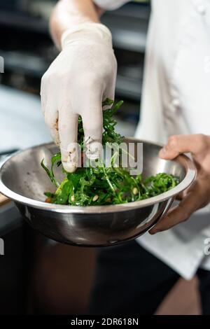 Gloved hand of contemporary chef of restaurant mixing chopped ruccola and beans in metallic bowl before putting the mixture into garnish Stock Photo