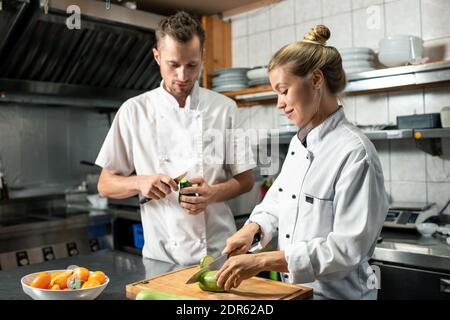 Young confident professional chef with knife peeling avocado and consulting his female trainee cutting fresh zucchini on wooden board Stock Photo