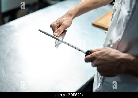 Hands of young male chef of modern restaurant in white uniform sharpening knife over kitchen table before chopping raw ingredients Stock Photo