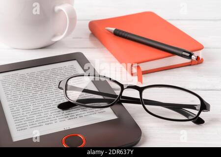 Eyeglasses on e-reader and orange note pad on a white wooden desk. Vision correction glasses for reading and computer work. Business and education. Stock Photo