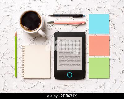 E-reader, cup of hot coffee, blank notepad, pensil, two pens and three colorful adhesive notes knolled on a white textured surface. Reading and writin Stock Photo