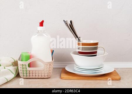 White dishwashing liquid and few foam sponges in a small basket near a stack of clean dishes on the kitchen table against white concrete wall. Stock Photo