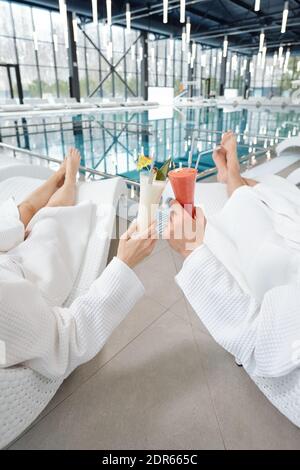 Hands of young relaxed man and woman in white bathrobes lying on deckchairs in front of camera and clinking with glasses of cocktails Stock Photo