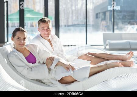 Young restful couple in white bathrobes sitting on deckchairs by poolside in front of camera at leisure center after swimming and looking at you Stock Photo
