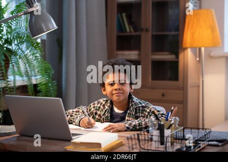 Clever and diligent African schoolboy making notes in copybook and looking at you while sitting by table in front of laptop in living-room Stock Photo