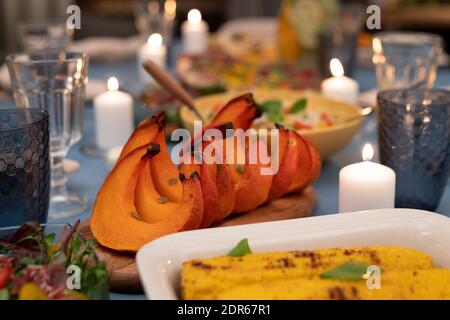 Part of served festive table with wineglasses, burning candles, vegetable salad, homemade grilled corn, baked pumpkin slices and other food Stock Photo