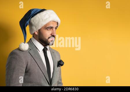 Charming bearded young man in Santa blue hat and grey suit, looking sideways isolated on yellow background. Pensive handsome emotions, facial Stock Photo