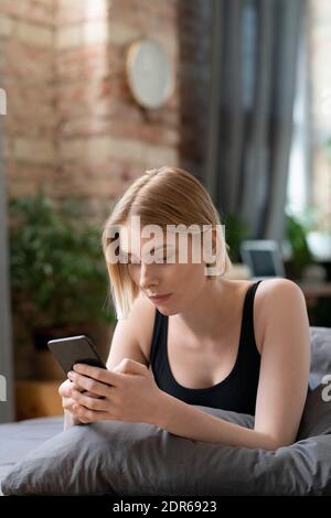 Young restful blond female with smartphone texting or scrolling through online news while relaxing on bed in bedroom on Sunday morning Stock Photo