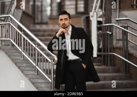 A young man in a black coat and white shirt posing outdoors. Handsome businessman looking aside and posing near the office stairs Stock Photo