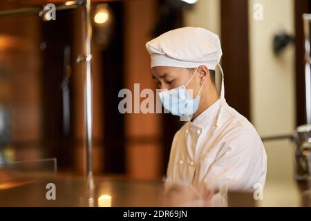 Side-view photo of attentive young chef wearing face mask while being focused on his work Stock Photo