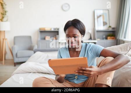 Minimal portrait of smiling African-American woman sitting on bed in morning and holding digital tablet, copy space Stock Photo
