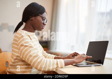 Side view portrait of young African-American woman using laptop while sitting at desk in home interior Stock Photo