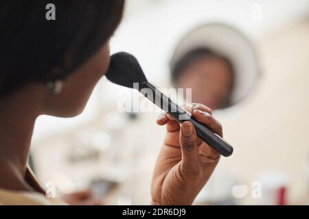 Close up of young African-American woman putting blush on cheeks with fluffy brush while doing makeup by mirror Stock Photo