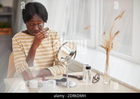 Portrait of young African-American woman using eyelash curler while doing makeup by mirror, copy space Stock Photo