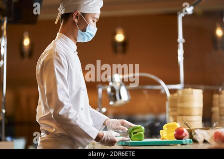 Side-view photo of a male Asian chef wearing protective face mask while cutting green pepper in his kitchen Stock Photo