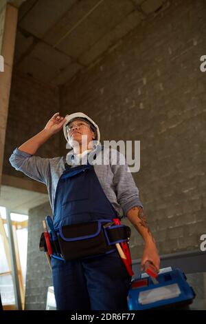 Low angle view of female construction worker wearing hardhat and holding toolbox while standing in concrete building Stock Photo