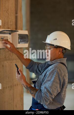 Vertical side view portrait of female worker setting up electricity and using smartphone while working on construction site Stock Photo