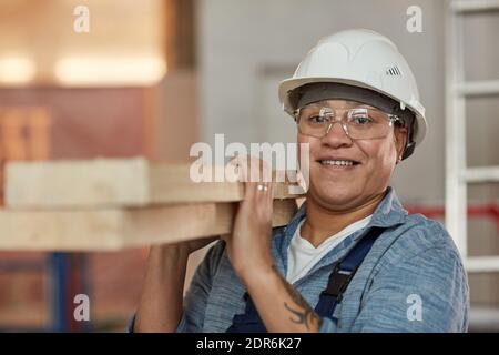 Front view portrait of smiling female worker carrying wood boards while working on construction site, copy space Stock Photo