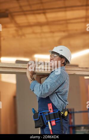 Vertical side view portrait of modern female worker carrying wood boards while working on construction site Stock Photo