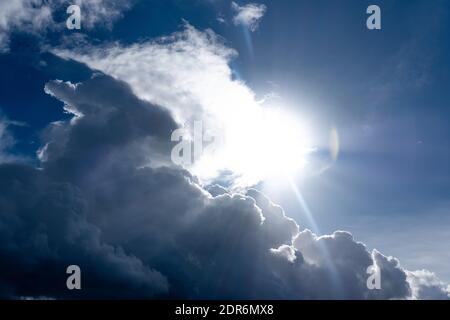 Natural sky background. Grey and white fluffy clouds hanging on the blue sky. Sun shines bright behind the clouds. Stock Photo