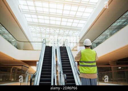 Back view portrait of female engineer holding blueprints while standing by escalators in shopping mall, copy space Stock Photo