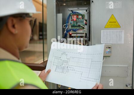 Female worker holding blueprints while inspecting electrical switchboard at construction site, copy space Stock Photo
