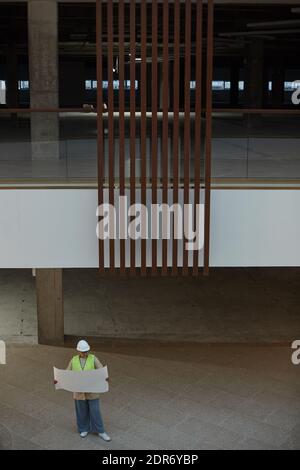 Vertical wide angle view at female engineer holding blueprints while standing in shopping mall or office building under construction, copy space Stock Photo