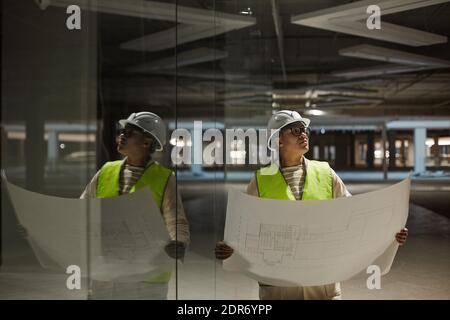 Waist up portrait of female engineer holding blueprints while inspecting base of building at construction site, copy space Stock Photo