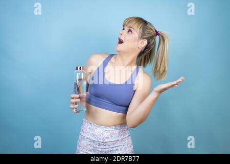 Young beautiful woman wearing sportswear holding a bottle of water over isolated blue background scared with her arms up like something falling from a Stock Photo