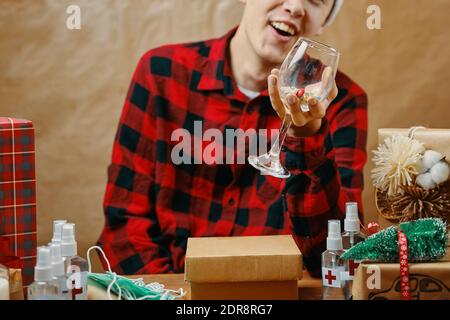 Guy in plaid shirt holds a glass with drugs and vitamins. Man in Christmas decorations. Table with gift boxes, antiseptics, medical masks and sweets. New year in quarantine. Festive atmosphere. Stock Photo