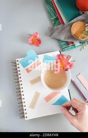 Empty spiral notebook full of multicolor reminder notes and a foamy coffee with a flower over it on a clean background. A woman hand is taking the gim Stock Photo