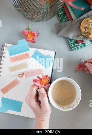 Woman left hand holding a foamy coffee over a notepad full of reminder notes and her right hand is holding a pencil ready to write. Stock Photo