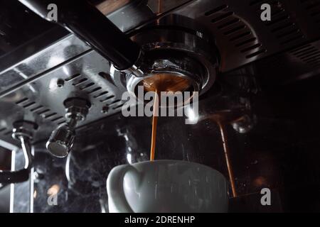 Preparation of espresso. Coffee pours from the coffee machine into the white cup. Professional coffee brewing. Stock Photo