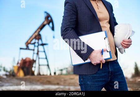 Close up of oil businessman holding protective helmet, clipboard, pen. Oil man standing on territory of oil field with pump jack on blurred background. Concept of petroleum industry and oil production Stock Photo