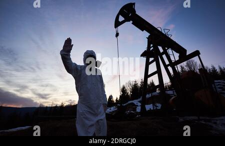 Scientist showing stop sign while standing on territory of oil field with pump jack, wearing protective suit, gas mask and gloves. Concept of ecology, petroleum industry and environmental pollution. Stock Photo