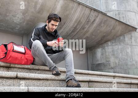 Delivery man with red bag uses his mobile phone to place next order, grey background Stock Photo