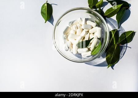 Capsules Vitamins and minerals in a glass bowl and a scattering of green leaves on a white background. Copy space for text Stock Photo
