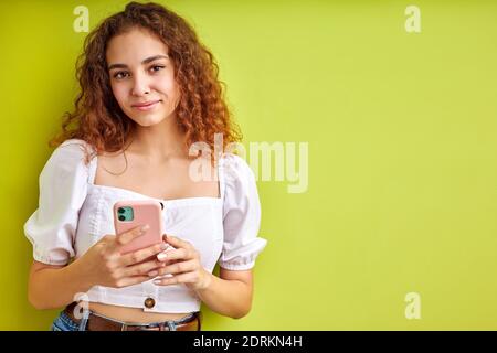 cute curly teen girl enjoy chatting with someone using mobile phone, adorable girl in blouse hold phone in hands, smile, isolated on green background Stock Photo