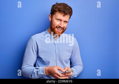 open-minded male is chatting with friend on smartphone, typing message, wearing blue formal shirt, smiling. isolated on blue background Stock Photo