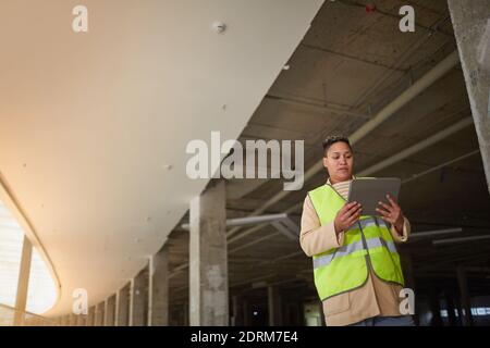Graphic wide angle portrait of mixed-race female engineer using digital tablet while standing on construction site, copy space Stock Photo