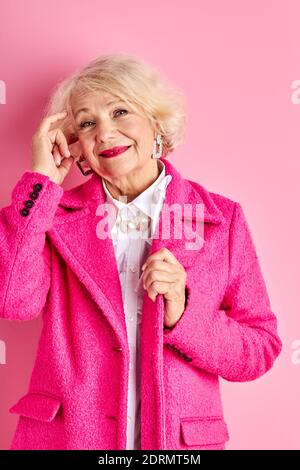 senior woman in stylish wear, beautiful grey haired lady enjoy her age, wearing fancy clothes, stand posing isolated over pink background Stock Photo