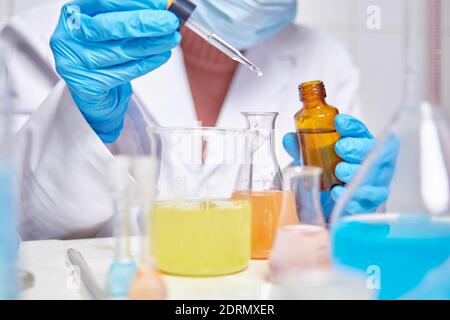 lab technician in white coat, blue gloves and face mask doing experiments and tests in a laboratory with different colored substances Stock Photo