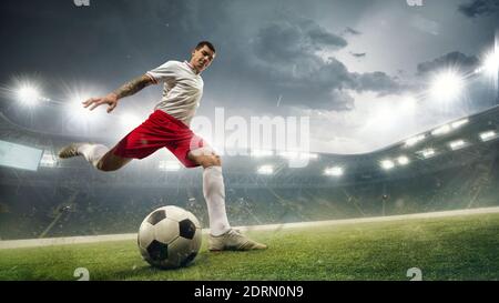 Young male soccer or football player in white and red sportswear kicking ball at the stadium in flashlights, spotlights. Concept of professional sport, motion, movement. 3D render. Flyer for ad. Stock Photo