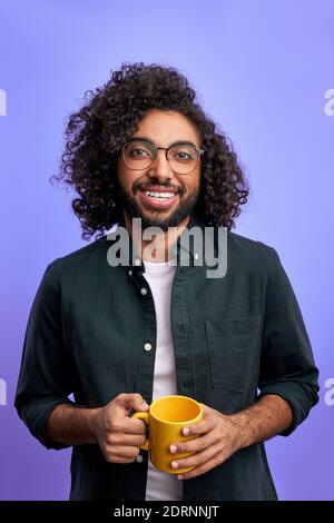 optimistic curly male begins the day with cup of tea of coffee, he is smiling at camera, wearing casual clothes, positive man isolated over purple bac Stock Photo