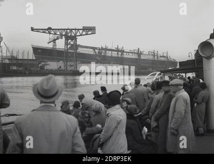 1930s, historical, passengers on a ship, looking across the harbour to a giant new ship being built, the RMS Queen Elizabeth, on the river Clyde, Glasgow, Scotland. UK. Built at the John Brown shipyard at Clydebank, for the Cunard White Star Line, she was the largest ship ever built at the time. Officially launched September 1938, she first entered service as a troopship during WWII, before taking passengers and mail on the Atlantic crossing until 1968. Stock Photo