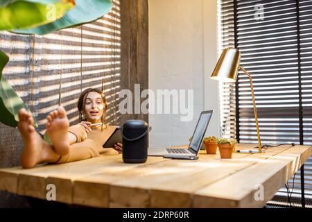 Woman talking to the smart speaker while working on the digital tablet at cozy home office. Concept of a smart home and Artificial Intelligence Stock Photo