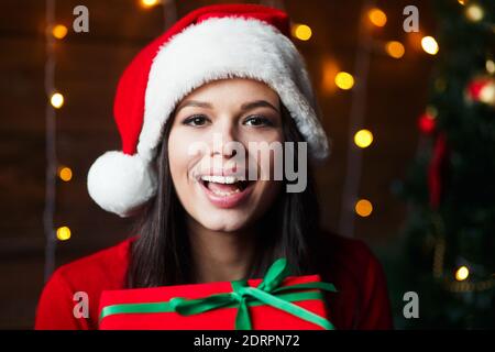 Happy young woman in Santa hat holding christmas gift at home Stock Photo