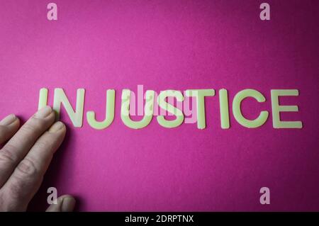 Human fingers holding the word Injustice, written with plastic letters on a bright pink paper background, concept Stock Photo