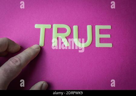 Human finger pointing the word True written with plastic letters on a bright pink paper background, concept Stock Photo